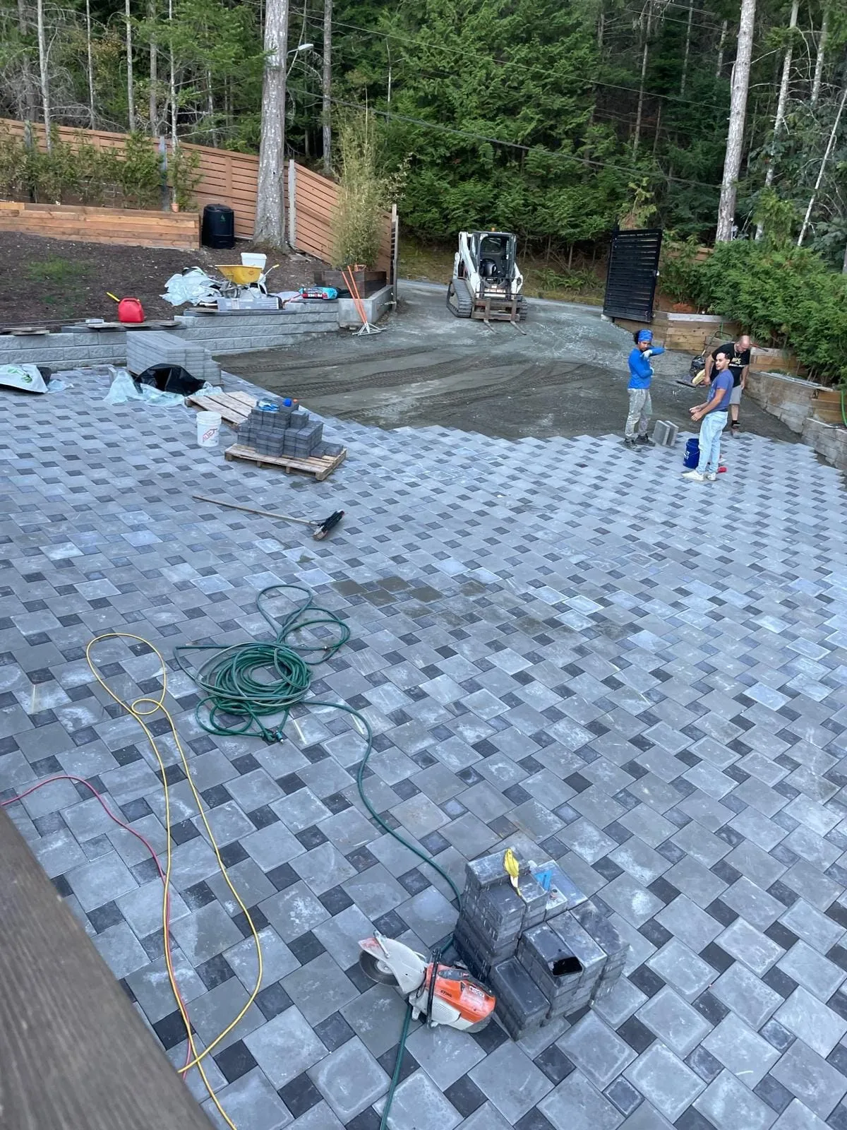 A wide paver field laid in a grey and charcoal pattern across a back yard, a cut off saw and stacked pavers in the foreground and three crew working along the far edge.