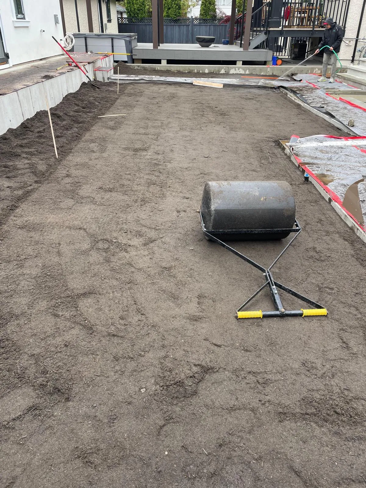 A long back yard bed of graded soil running up to a finished concrete patio, with a lawn roller in the foreground and a crew member watering the soil down at the far end.